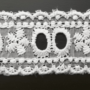 A close-up view of a roll of white cotton cambric lace trim with a detailed floral eyelet pattern.